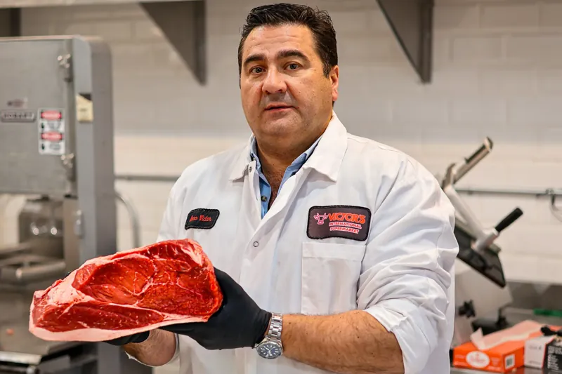 Nuno, con bata de Victor’s International Supermarket, sostiene un gran corte de carne de res cruda en la carnicería